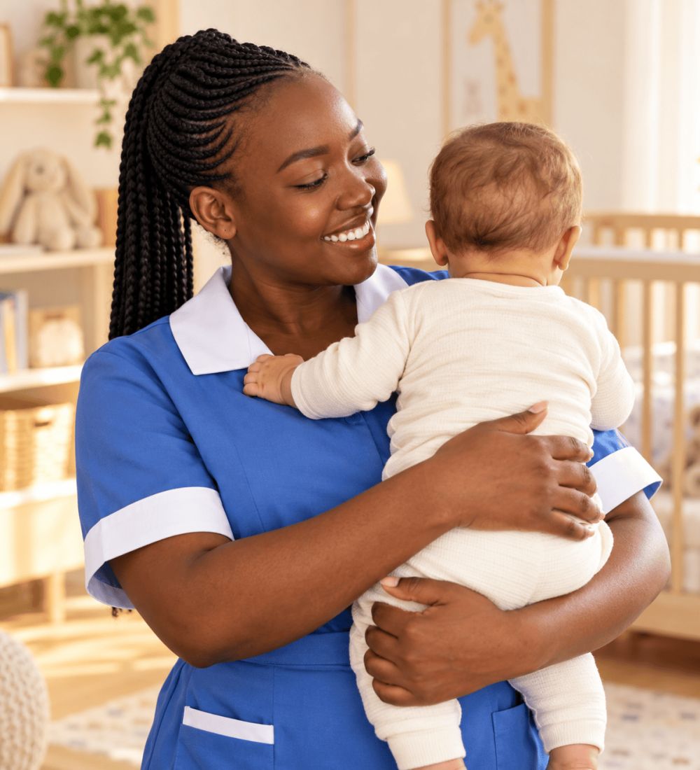 A smiling Domestic Angels nanny in a blue uniform holding a baby in a nursery